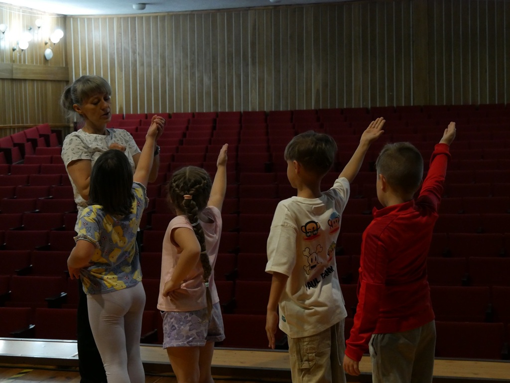 Kilkoro dzieci i kobieta zatrzymani w figurze tanecznej. W tle kilka rzędów czerwonych foteli.