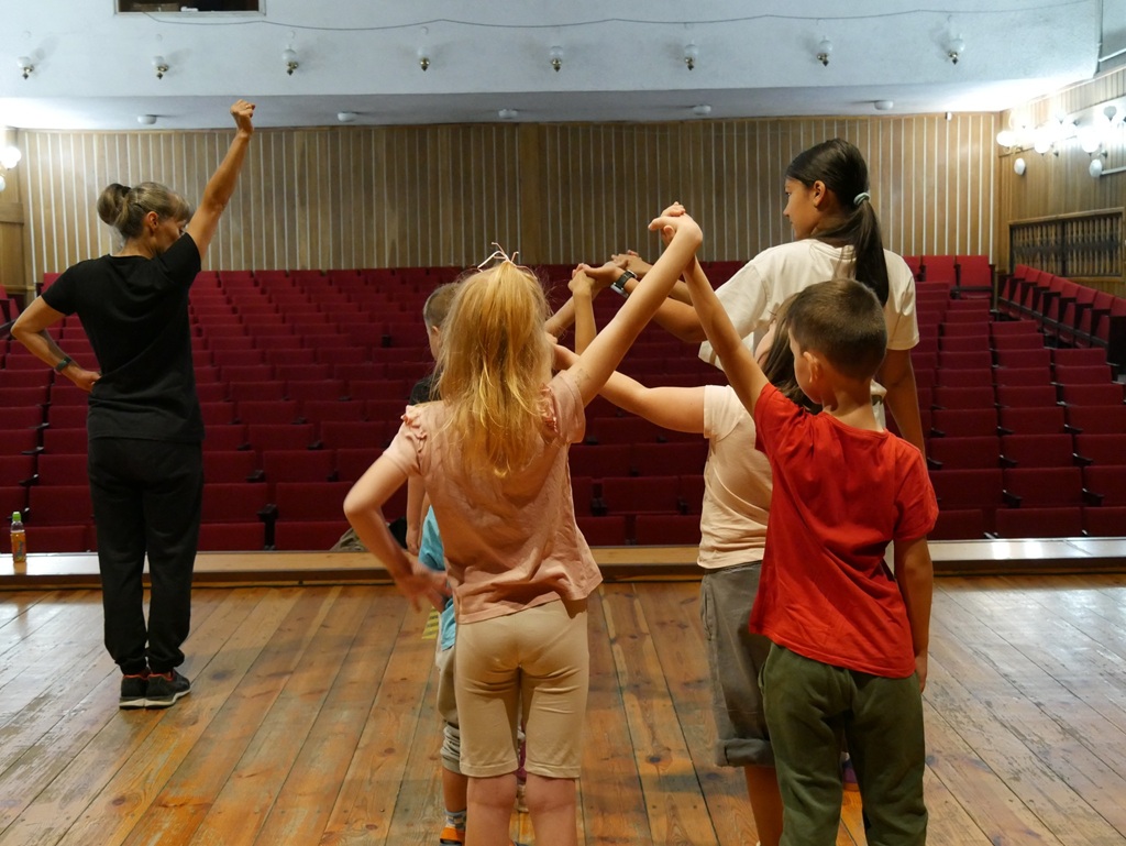 Kilkoro dzieci i kobieta zatrzymani w figurze tanecznej. W tle kilka rzędów czerwonych foteli. Pomieszczenie ciepło oświetlone.