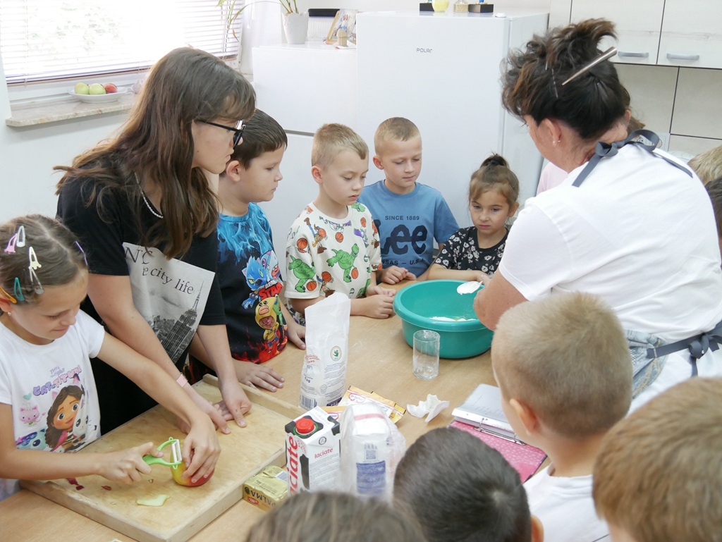 Dzieci i kobieta stoją przy stole, na którym stoją przybory i produkty kuchenne.