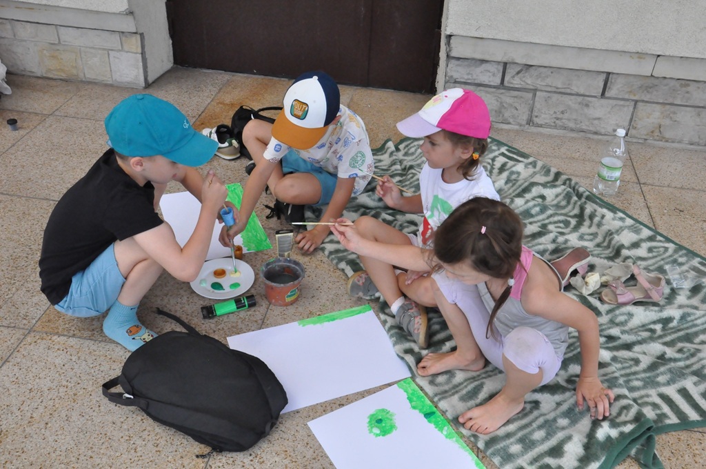 Dzieci siedzą na kocu i malują farbami na białych kartkach.