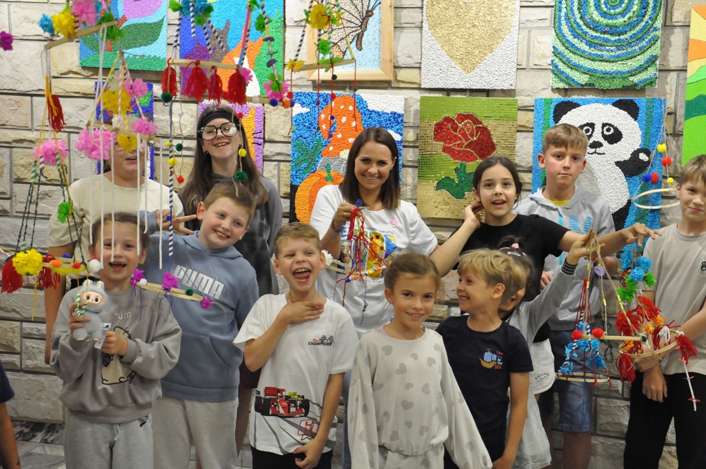 Dzieci i kobieta pokazują folklorystyczne pająki. Za nimi ściana z kolorowymi pracami. pomieszczenie ciepło oświetlone.