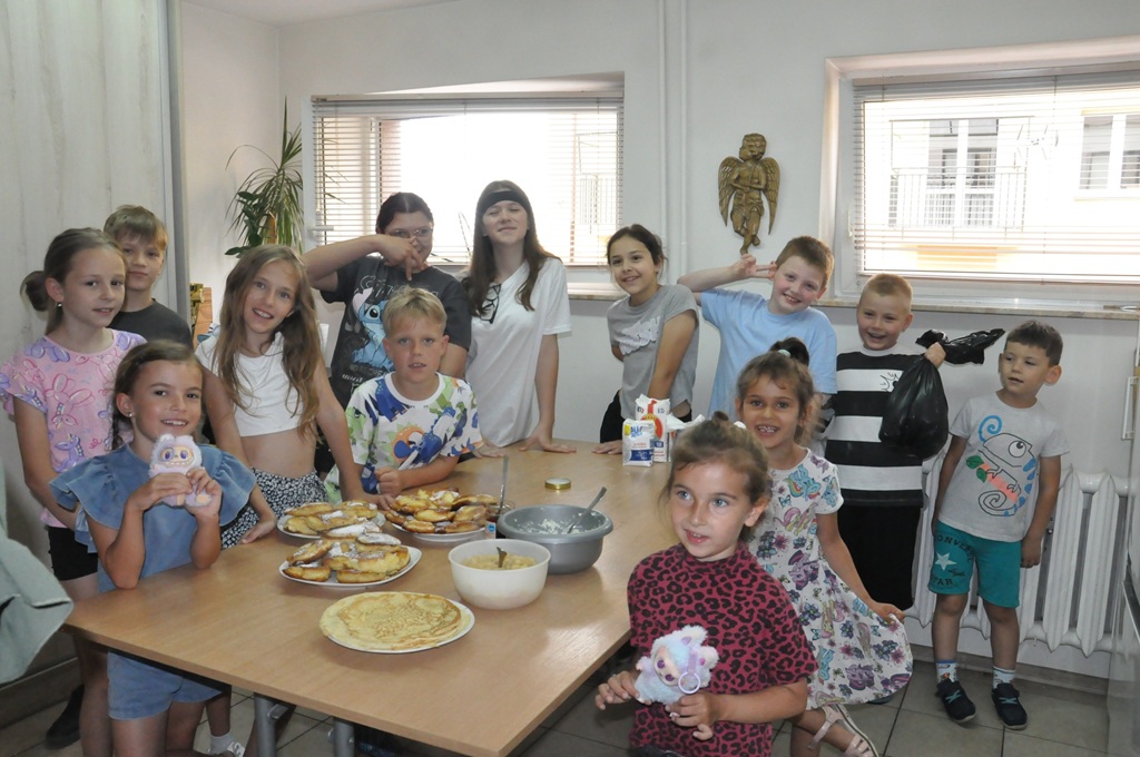Dzieci stoją lub siedzą przy stole, na którym leżą talerze z naleśnikami.
