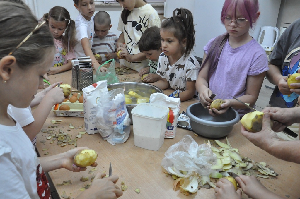 Dzieci w letnich ubraniach siedzą lub stoją przy stole z produktami i przyborami kuchennymi. Pomieszczenie ciepło oświetlone.