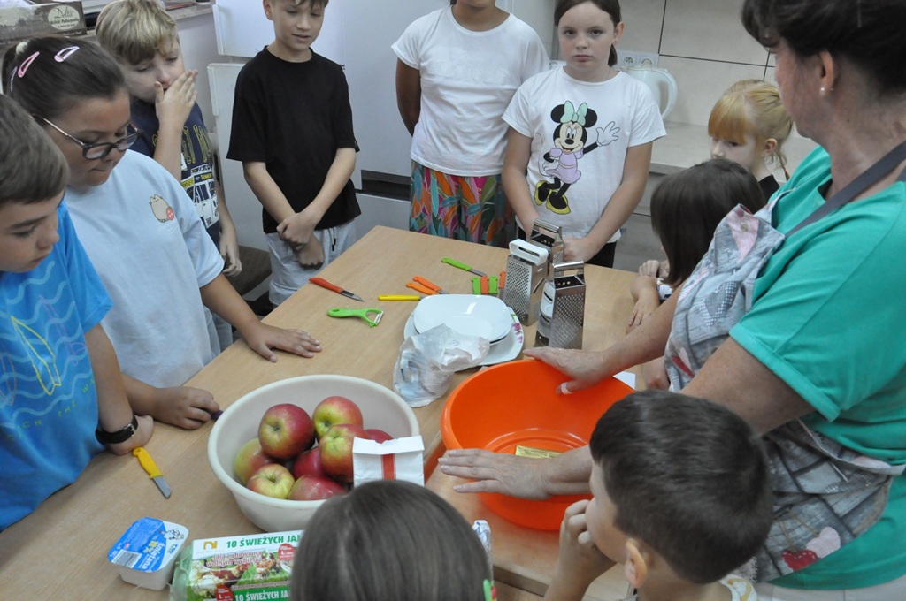 Dzieci w letnich ubraniach i kobieta w fartuchu siedzą lub stoją przy stole z produktami i przyborami kuchennymi. Pomieszczenie ciepło oświetlone.