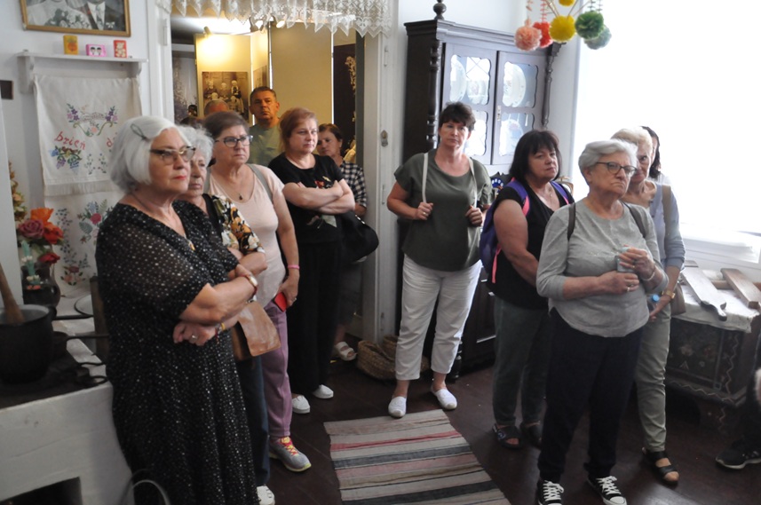 Grupa dorosłych osób w letnich ubraniach ogląda wiejską chatę.