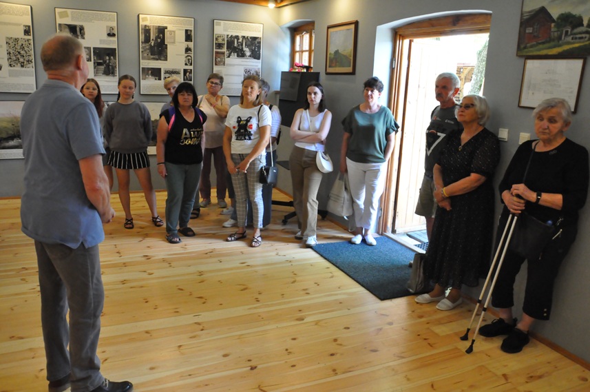 Grupa dorosłych osób ogląda ekspozycję chaty ludowej. Pomieszczenie ciepło oświetlone.