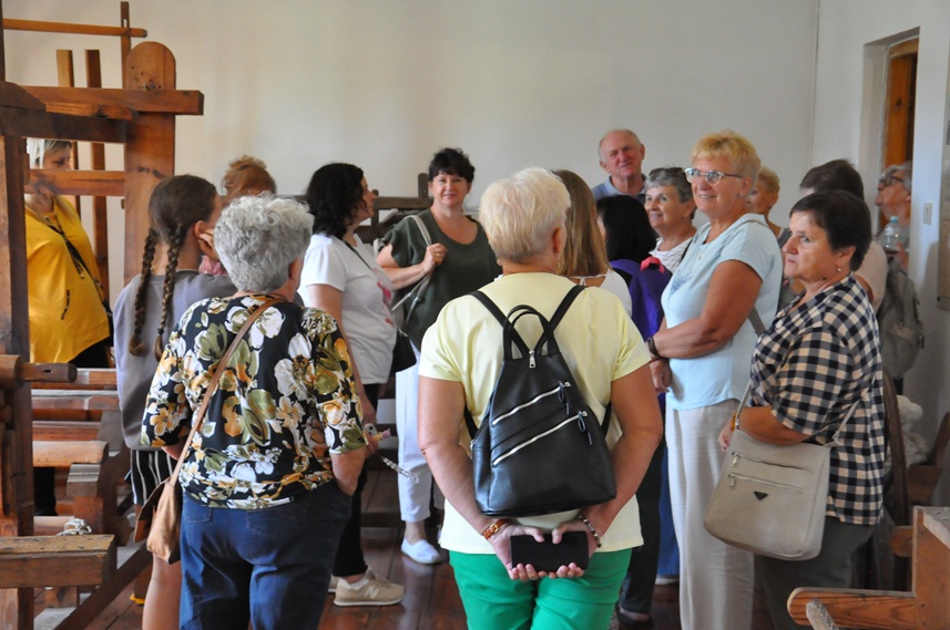 Grupa dorosłych osób w letnich ubraniach ogląda ekspozycję chaty ludowej.