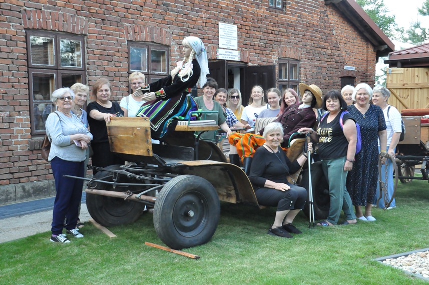 Duża grupa dorosłych osób pozuje do wspólnego zdjęcia przy szlacheckim wozie. W tle budynek z czerwonej cegły. Pogodny dzień.