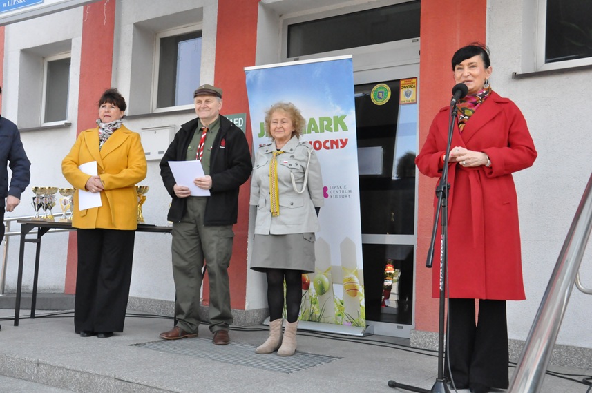 Kobieta w czerwonym płaszczu stoi przy mikrofonie na statywie. Obok niej stoi mężczyzna i kobieta w harcerskich strojach, obok nich jest jeszcze jedna kobieta w żółtej marynarce.