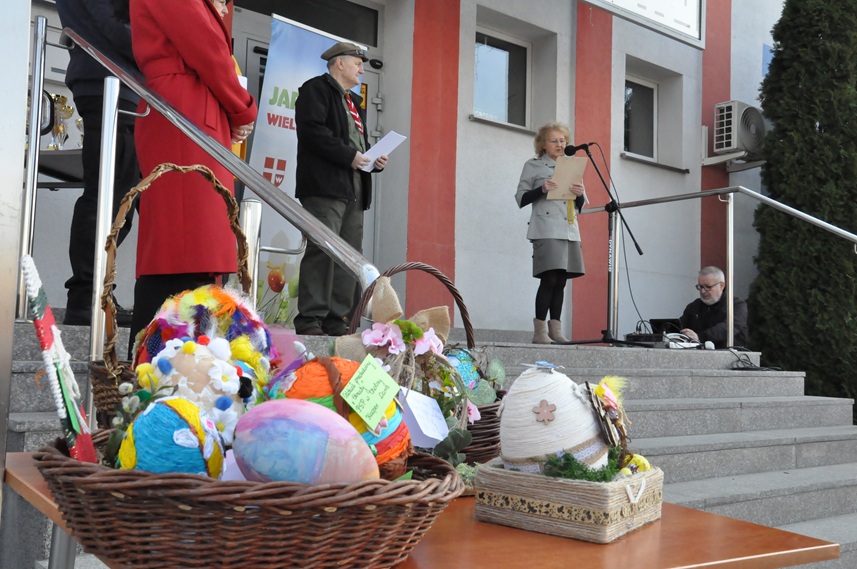 Kobieta w harcerskim stroju stoi przy mikrofonie na statywie. Obok niej stoi jeszcze kilka osób. Na pierwszym planie stół, na których leżą kolorowe pisanki.