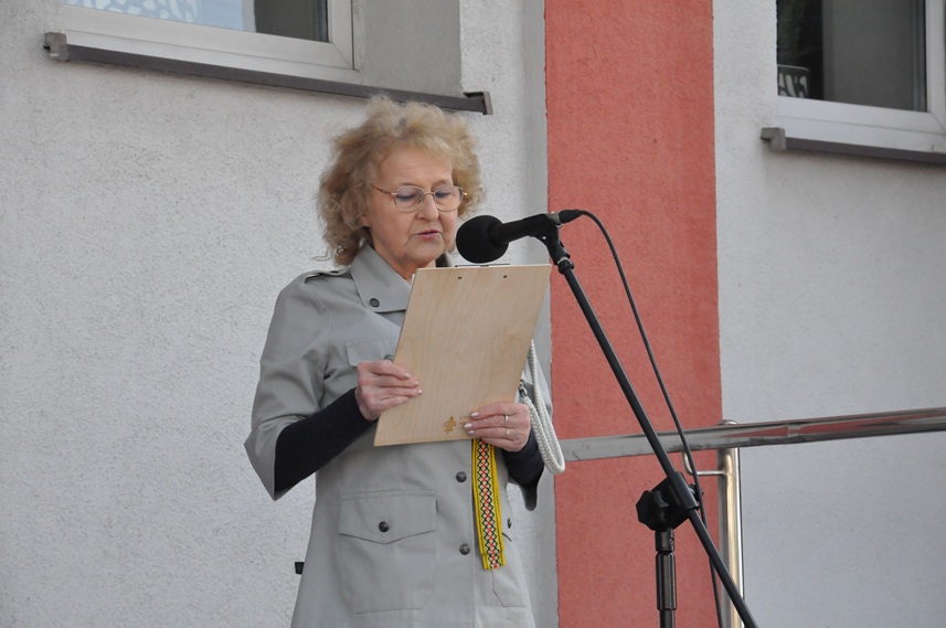 Kobieta w harcerskim stroju stoi przy mikrofonie na statywie. Za nią szara ściana budynku z oknami.