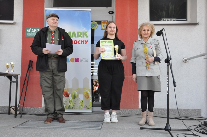 Dwoje dorosłych w harcerskich strojach i dziewczyna w czarnym ubraniu pozują do wspólnego zdjęcia. dziewczyna trzyma dyplom i puchar.