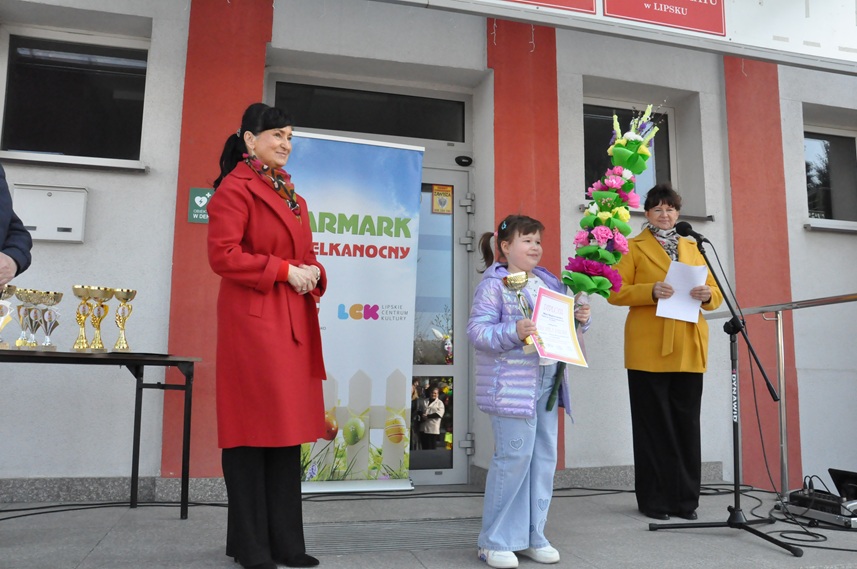 Dwie kobiety i dziewczynka pozują do wspólnego zdjęcia. Dziewczynka trzyma dyplom, puchar i palmę wielkanocną.