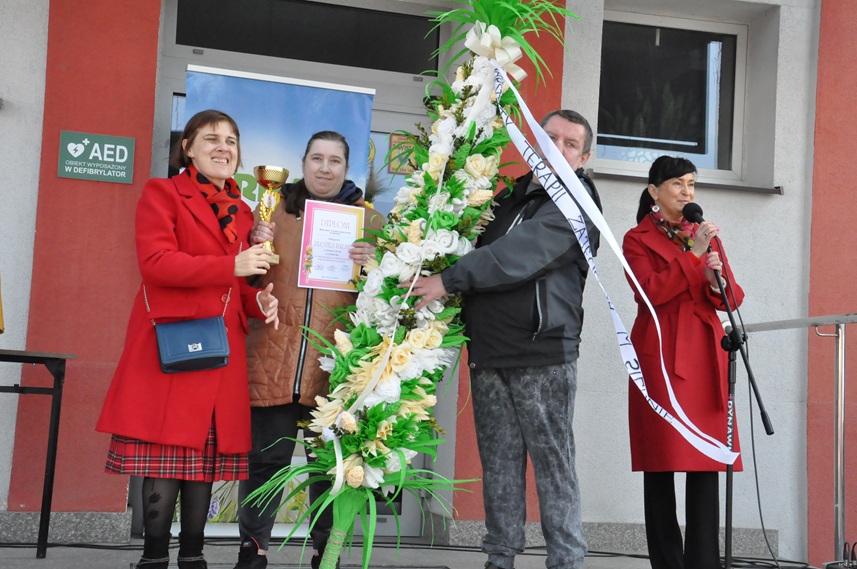 Cztery osoby pozują do wspólnego zdjęcia. Kobieta w czerwonym płaszczu stoi przy mikrofonie na statywie. Mężczyzna w czarnej kurtce trzyma dużą palmę wielkanocną. Kobieta w brązowej kurtce trzyma dyplom, a jeszcze jedna kobieta w czerwonym płaszczu puchar.