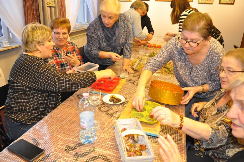 Kilka kobiet siedzi lub stoi przy stole. Wszystkie przygotowują dekoracyjne świece.