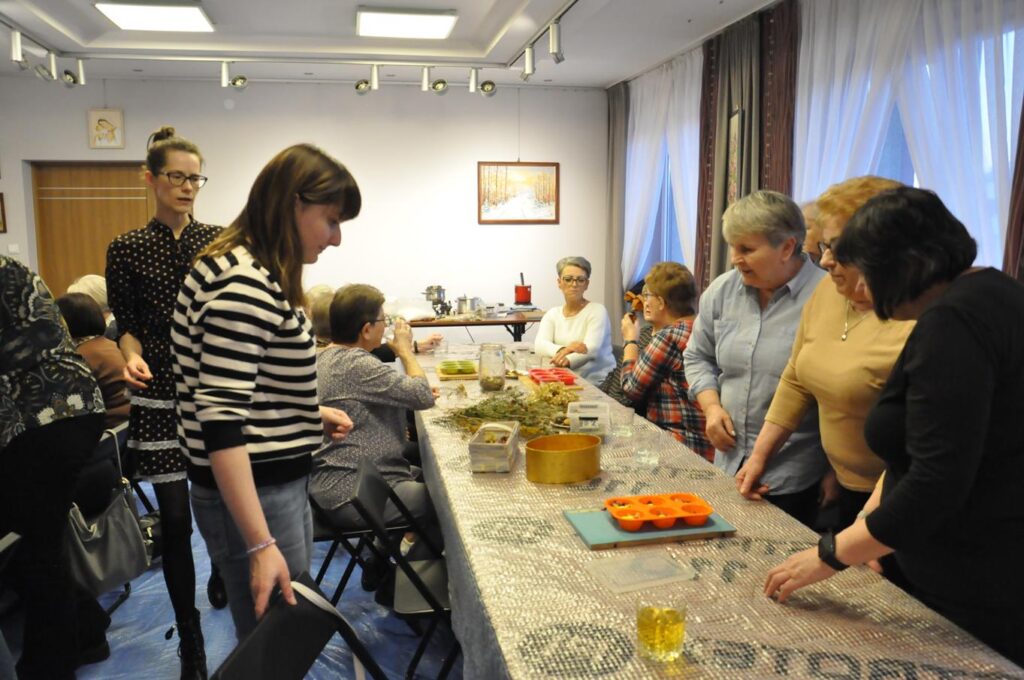Kilka kobiet przy stole. Niektóre siedzą, niektóre stoją. Na stole przybory do stworzenia świec. Pomieszczenie ciepło oświetlone.