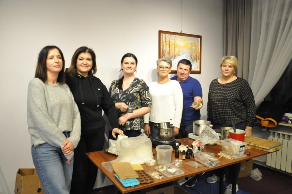 Kilka kobiet i mężczyzna w granatowej koszulce pozują do wspólnego zdjęcia przy stole z przyborami do stworzenia aromatycznych świec. Pomieszczenie ciepło oświetlone.