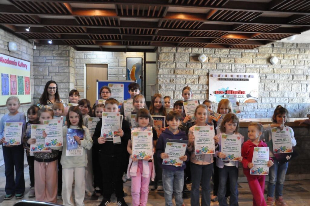 Dzieci pozują do wspólnego zdjęcia Wszystkie trzymają pamiątkowe dyplomy i nagrody. Pomieszczenie ciepło oświetlone.