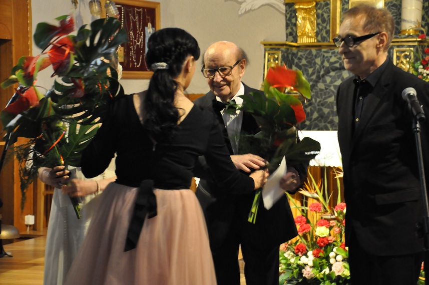 Kobieta w czarnej bluzce i różowej sukience podaje bukiet kwiatów mężczyźnie w czarnym garniturze. Obok stoi kolejny mężczyzna w czarnym garniturze. Za nimi ołtarz kościelny.