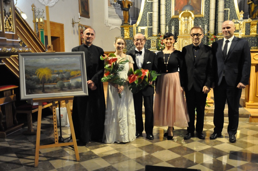 Grupa sześciu osób, mężczyzn i kobiet w galowych ubraniach oraz ksiądz stoją przy obrazie wyeksponowanym na stelażu. Obok nich ambona. Za nimi ołtarz kościelny.