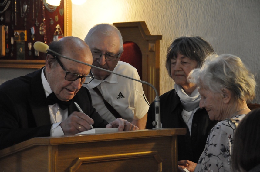 Cztery osoby w wieku senioralnym. Troje z nich patrzy na to co czwarta osoba, mężczyzna w okularach notuje. W tle tablica z wisiorami.