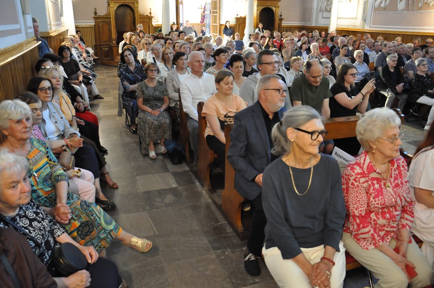 Duża grupa ludzi siedzi w kościelnych ławach. Za nimi dwa konfesjonały.