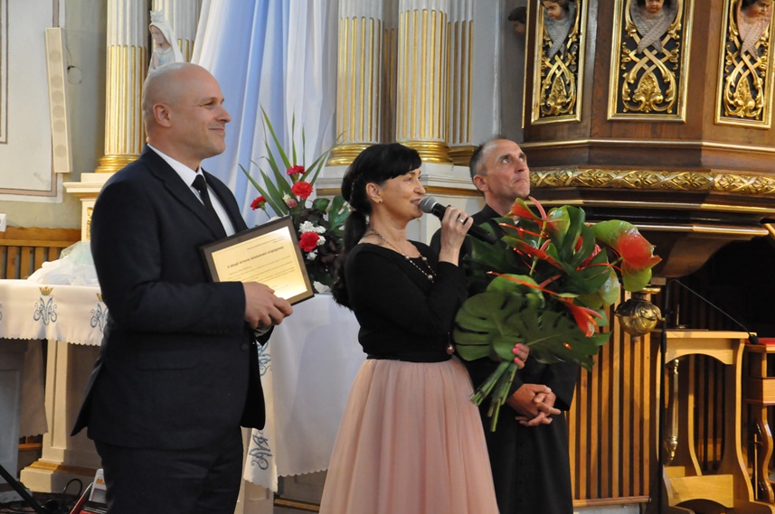 Kobieta w czarnej bluzce i różowej sukience trzyma mikrofon przy ustach, trzyma bukiet kwiatów i stoi między księdzem, a mężczyzną w czarnym garniturze który trzyma grawerton. Za nimi kościelna ambona.