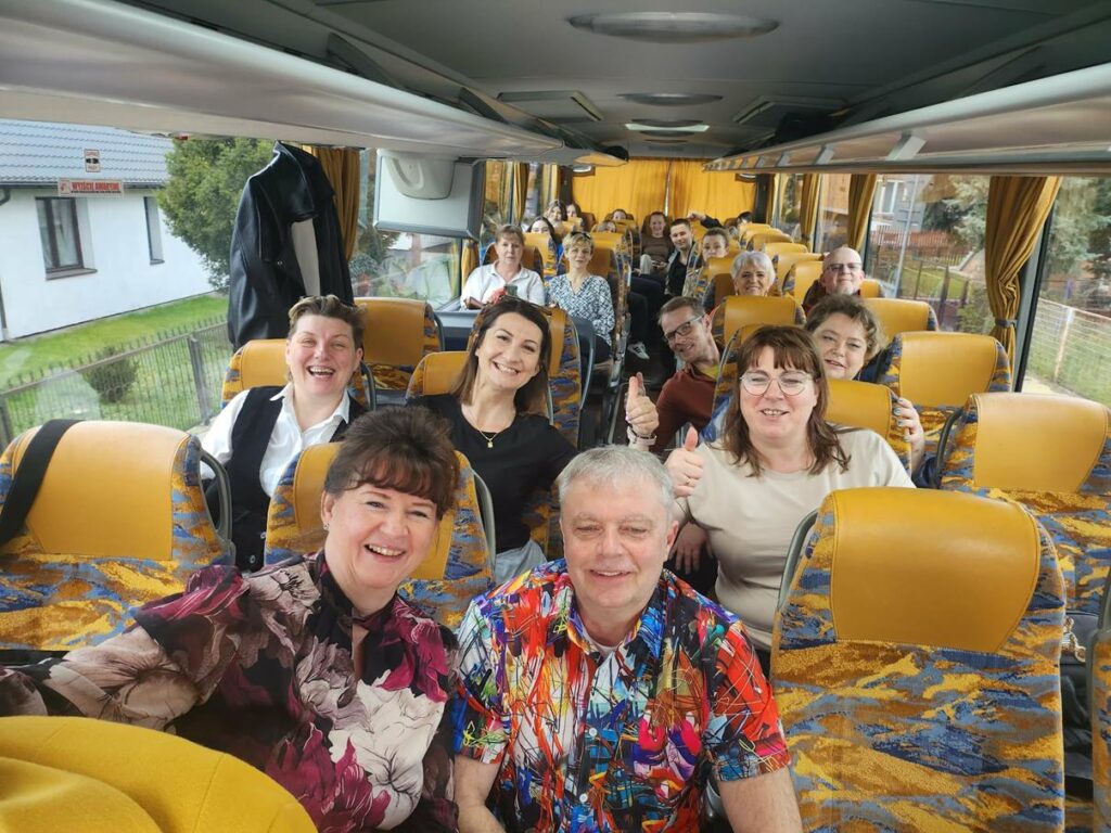 Grupa dorosłych osób siedzi na fotelach w autobusie. Wszyscy się uśmiechają, niektórzy pokazują kciuki do góry.