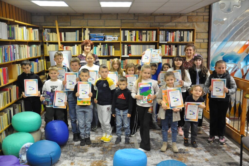Duża grupa dzieci i dwie kobiety pozują do wspólnego zdjęcia. Wszyscy stoją. Dzieci trzymają dyplomy i książki. Za nimi regały z książkami. Pomieszczenie ciepło oświetlone.