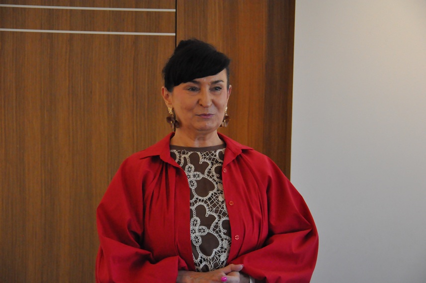 Kobieta w czerwonej koszuli z ujętymi przed sobą dłońmi. Pomieszczenie ciepło oświetlone.