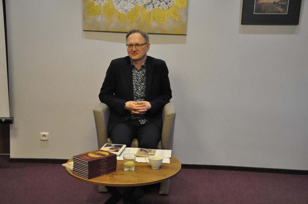 Mężczyzna w czarnej marynarce i w okularach siedzi przy niskim okrągłym stoliku na którym leżą książki, filiżanka i szklanka z wodą. Za nim szara ścina z obrazami.