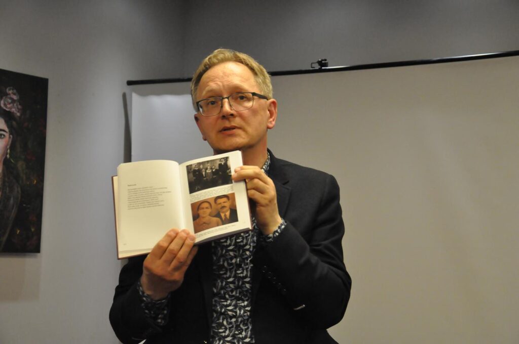Mężczyzna w czarnej marynarce i w okularach pokazuje stare fotografie w książce.