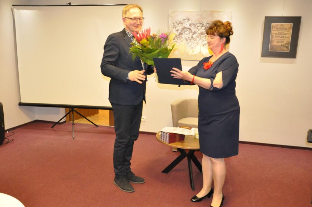 Kobieta w grafitowej sukience podaje mężczyźnie w czarnej marynarce i w okularach granatową teczkę. Mężczyzna trzyma bukiet kolorowych kwiatów. Za nimi duży ekran na statywie oraz ściana z obrazami.
