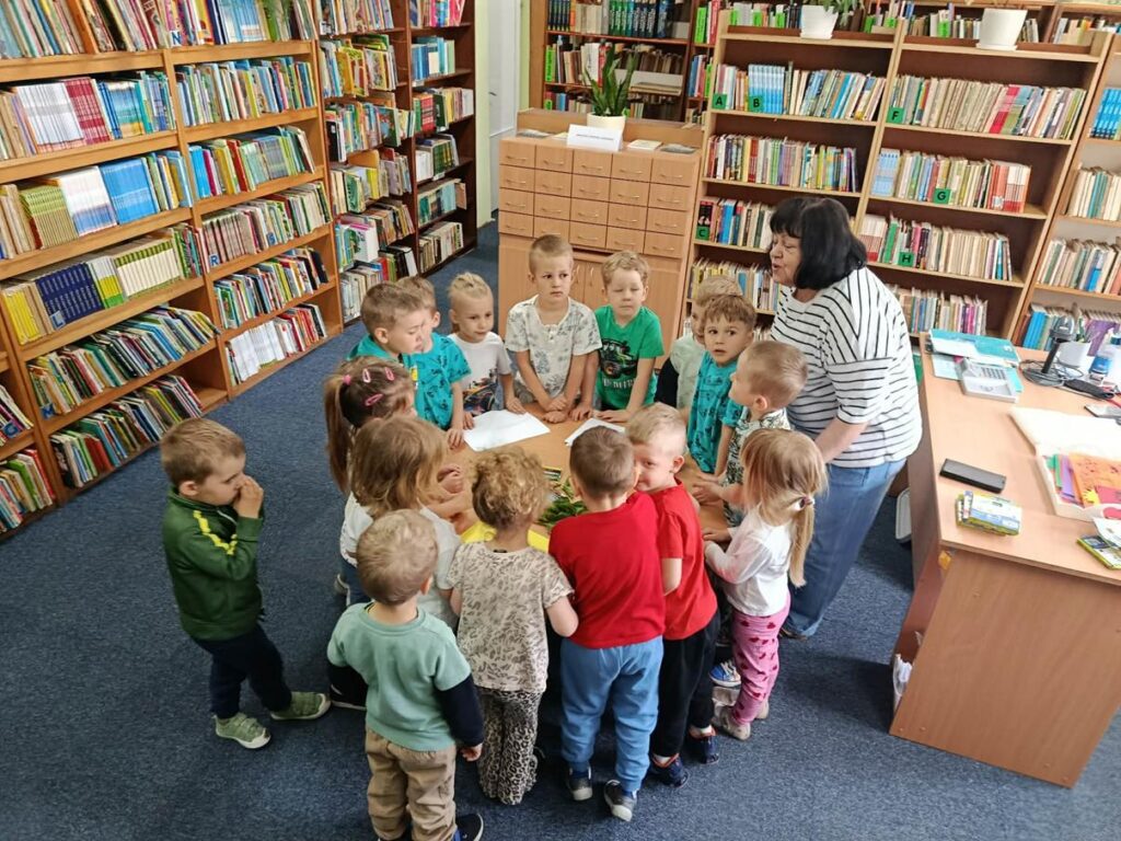 Grupa dzieci i kobieta stoją wokół okrągłego stołu. Wokół regały z książkami. Pomieszczenie ciepło oświetlone.