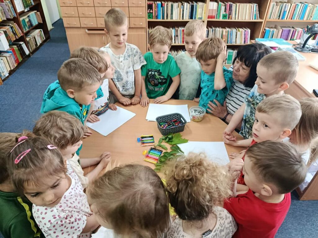 Grupa dzieci zgromadzona wokół okrągłego stołu na którym zgromadzone są kartki białego papieru, kolorowe kredki i liście. Wokół regały z książkami. pomieszczenie ciepło oświetlone.