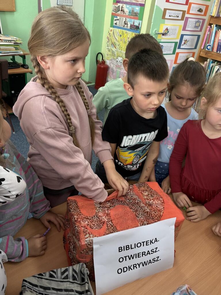 Dziewczynka wkłada rękę do pudełka z pomarańczowym wzorem. Obok niej stoi więcej dzieci. Za nimi zielona ściana z kolorowymi obrazkami.