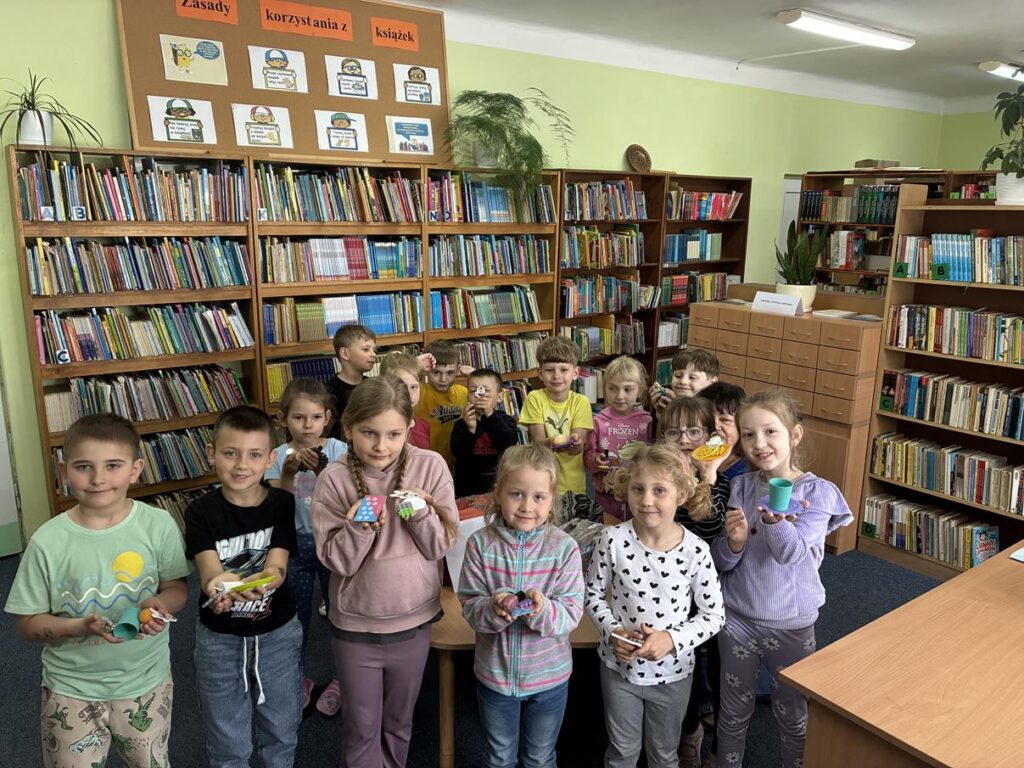 Grupa dzieci stoi w bibliotece. Trzymają różne kolorowe przedmioty. Pomieszczenie ciepło oświetlone.