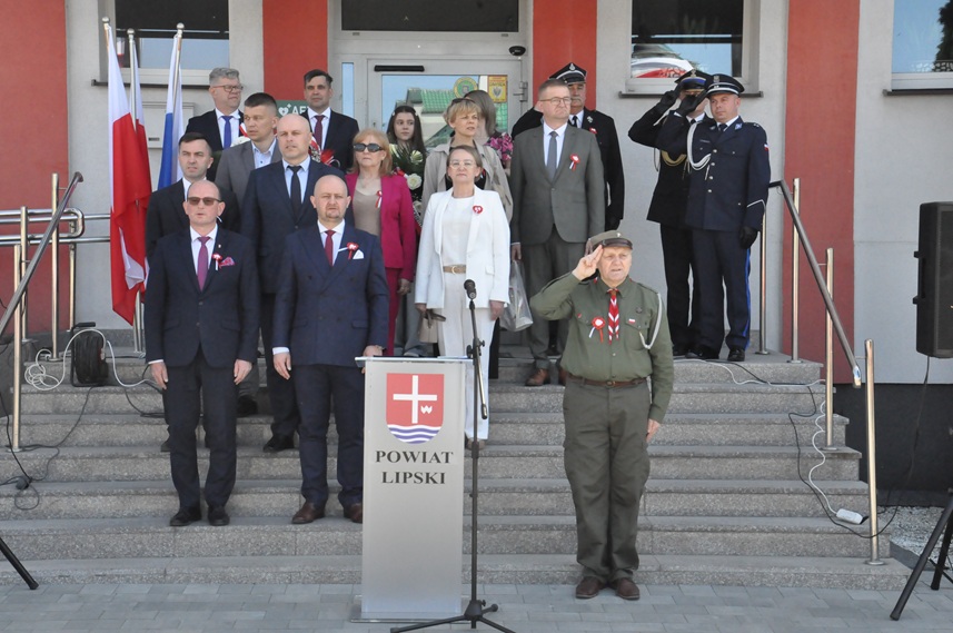 Grupa dorosłych osób stoi na schodach w odświętnych ubraniach. Niektórzy salutują.