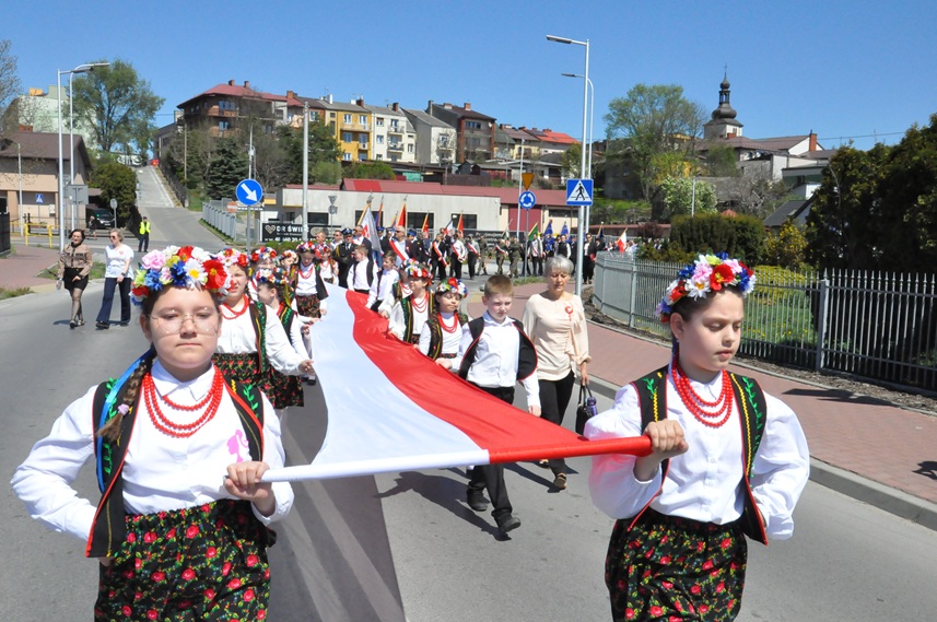 Dzieci w ludowych strojach niosą ogromną flagę z barwami narodowymi. Dziewczynki mają warkocze i kolorowe wianki na głowach. Za nimi panorama miasteczka z wieżą kościelną.
