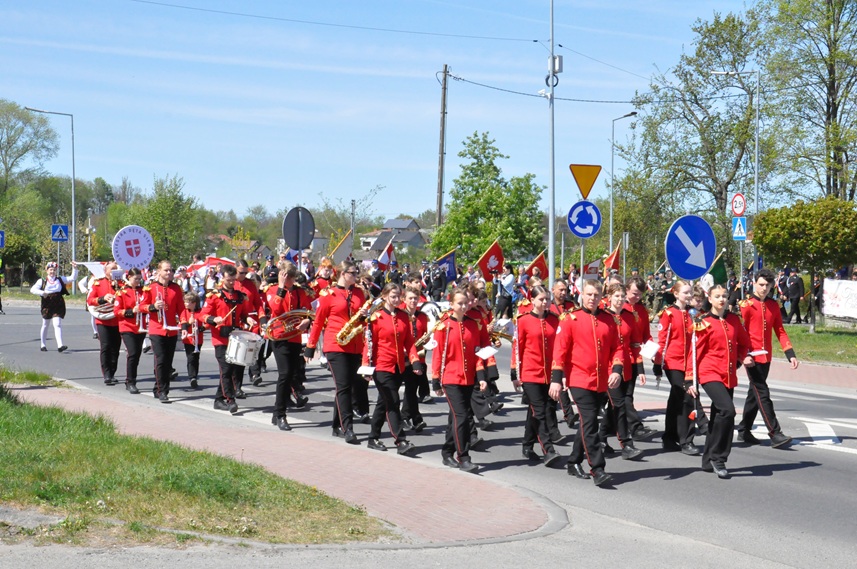 Orkiestra dęta w czerwonych strojach maszeruje ulicą. Pogodny dzień.