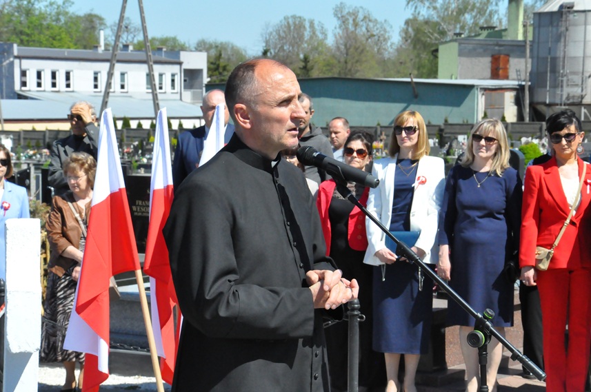 Ksiądz stoi przy mikrofonie na statywie. Za nim flagi Polski i kilka osób w odświętnych strojach. Pogodny dzień.