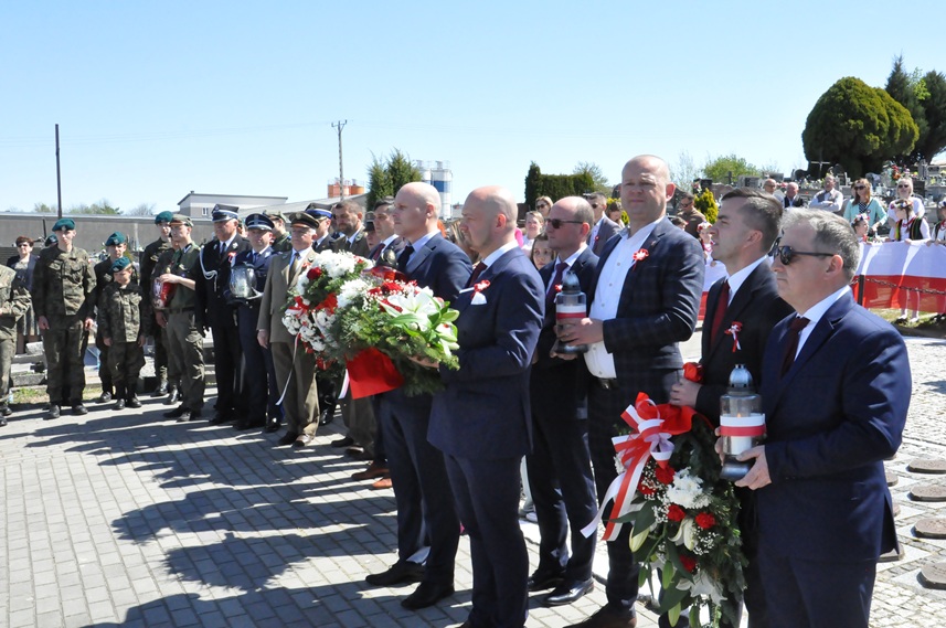 Mężczyźni w garniturach trzymają znicze i wiązanki kwiatów w barwach narodowych. Za nimi mężczyźni i młodzież w mundurach. pogodny dzień.