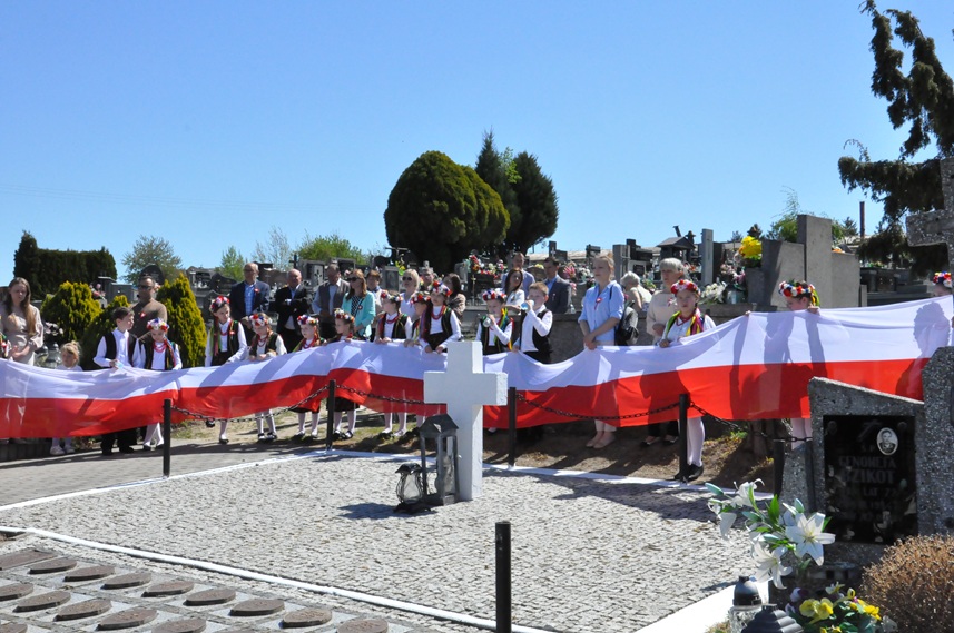 Dzieci trzymają ogromną flagę w barwach narodowych. Wokół stoją dorośli. Za nimi są nagrobki i kilka drzew. Pogodny dzień.