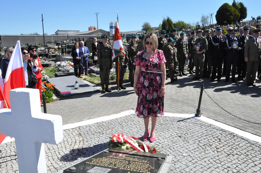 Kobieta w sukience w różowe kwiaty składa hołd przed pomnikiem. Za nią stoją osoby w mundurach i odświętnych strojach. pogodny dzień
