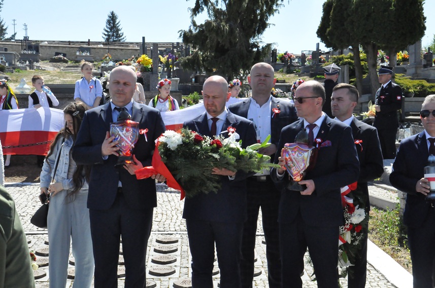 Kilku mężczyzn w garniturach stoi i trzyma znicze oraz wiązanki z kwiatów o barwach narodowych. Za nimi więcej osób, nagrobki i kilka drzew. Pogodny dzień.