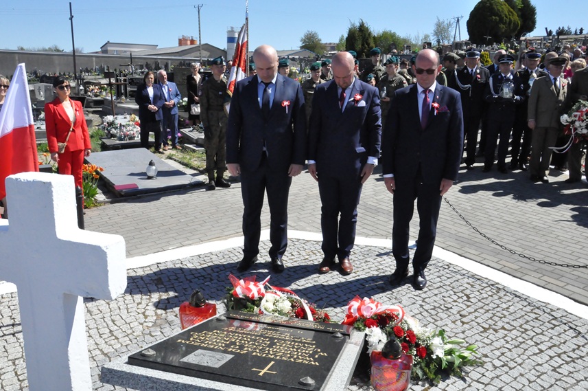 Trzech mężczyzn w grafitowych mundurach składa hołd przed pomnikiem. Za nimi stoją osoby w mundurach i odświętnych strojach. Pogodny dzień.