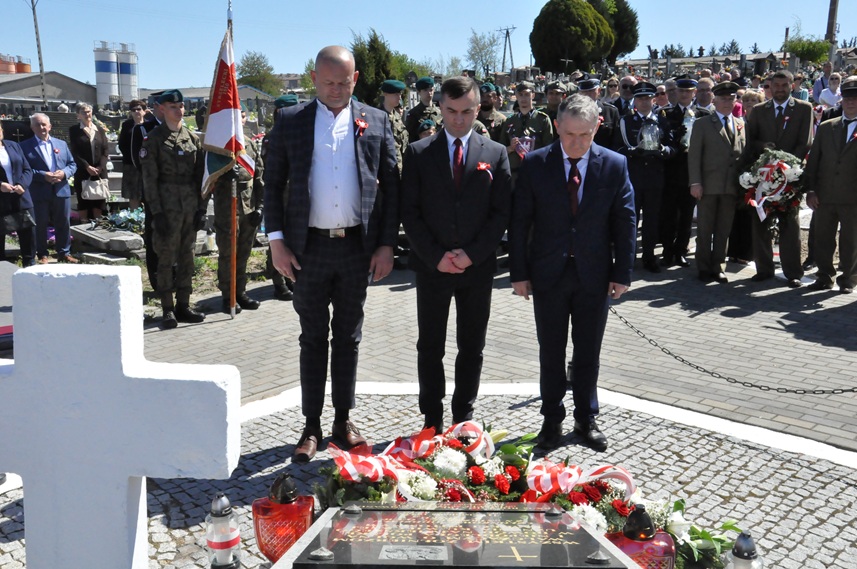 Trzech mężczyzn w garniturach składa hołd przed pomnikiem. Za nimi stoją osoby w mundurach i odświętnych strojach. Dalej nagrobki i kilka drzew. Pogodny dzień.