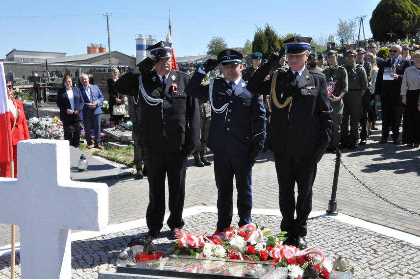Trzech mężczyzn w policyjnych i strażackich mundurach składa hołd przed pomnikiem. Za nimi stoją osoby w mundurach i odświętnych strojach. Dalej nagrobki i kilka drzew. Pogodny dzień.