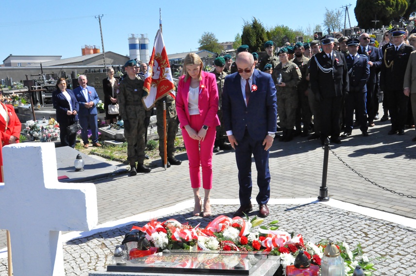 Kobieta w różowym stroju i mężczyzna w grafitowym garniturze składają hołd przed pomnikiem. Za nimi stoją osoby w mundurach i odświętnych strojach. Dalej nagrobki i kilka drzew. Pogodny dzień.