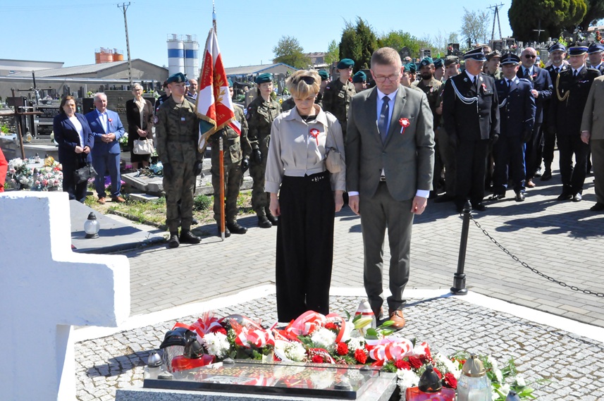Kobieta w szarej koszuli i czarnych spodniach oraz mężczyzna w jasnym garniturze składają hołd przed pomnikiem. Za nimi stoją osoby w mundurach i odświętnych strojach. Dalej nagrobki i kilka drzew. Pogodny dzień.